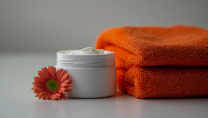 White round plastic jars filled with white cream, placed on a table next to two bright orange towels and a small delicate flower, set against a clean white background,