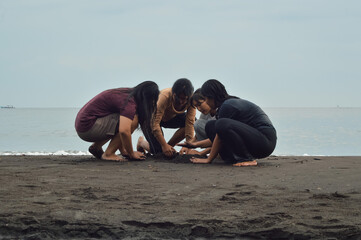 The girls huddle over wet sand as the tide laps nearby, sculpting shapes together while one remains...