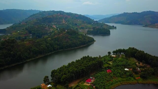 Drone view over Lake Bunyonyi in southwestern Uganda showing green peninsula with cultivated plots and scattered houses in the foreground, and forested terraced hills surrounding the calm lake waters.