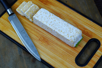 A block of tempeh, partially sliced, on a wooden cutting board with a knife, set against a woven bamboo background.