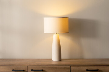 Tapered ceramic table lamp with drum linen shade, styled on a natural oak wood console with off-white stoneware base, set against a beige wall and illuminated by soft warm ambient light and shadows.