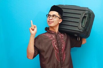 Portrait of surprised indonesian muslim man carrying suitcase while pointing finger with smiling happy expression. Going home for Eid Mubarak or Mudik Isolated image on blue background.