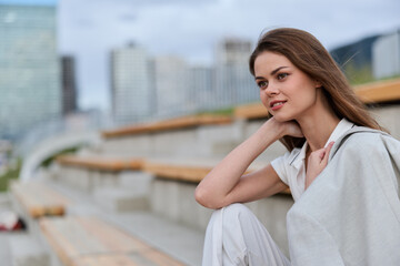 Fototapeta premium Young woman in white casual wear sitting thoughtfully outdoors with cityscape background, natural light and calm expression in spring fashion style