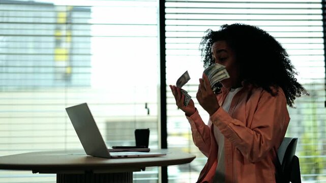 Excited African American woman in pink shirt and white top counting money at desk in modern office. She beams with joy, celebrating financial success and prosperity, achieving her goals.