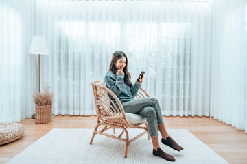 Young Asian Woman Relaxing in Modern Wicker Chair Near Window with Sheer Curtain, Happy Female...