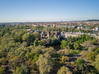 Fototapeta premium Wider aerial photo of Vajdahunyad Castle and artificial lake in Budapest, centered in lush City Park surroundings, with expanded view of greenery and park paths in soft morning light. 