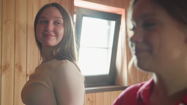 sisters exchange warm smiles while standing in wooden room near window, sharing lighthearted joyful moment together just before falling backward on bed