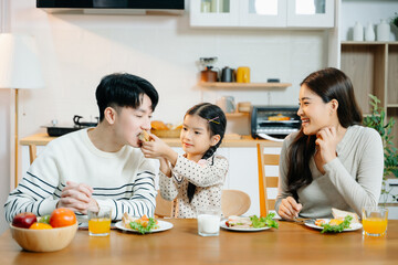 Asian father reads book to daughter at breakfast while mom prepares food. Heartwarming family...