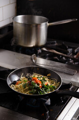 A frying pan filled with spaghetti, fresh spinach, and cherry tomatoes is cooking on a gas stove, with a saucepan of boiling water in the background of a professional kitchen