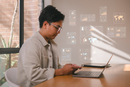 A young man uses his smartphone for messaging while working on a laptop, with digital data charts. Concept of remote work, social media, and mobile technology