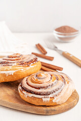 Homemade sweet freshly baked cinnamon rolls bun glazed with sugar icing served on wooden cutting board on white table with spice powder, sticks and fork for breakfast as pastry dessert comfort food