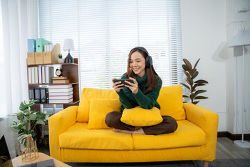 Obraz premium Leisure Play: A woman relaxes on a sunny yellow couch, engrossed in a handheld gaming session while wearing headphones, a portrait of comfort, leisure, and modern home entertainment.