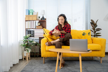 Fototapeta premium Cozy Lifestyle: A woman with headphones on relaxes and works on a laptop on a yellow sofa, enjoying leisure time, soft light and a modern atmosphere.