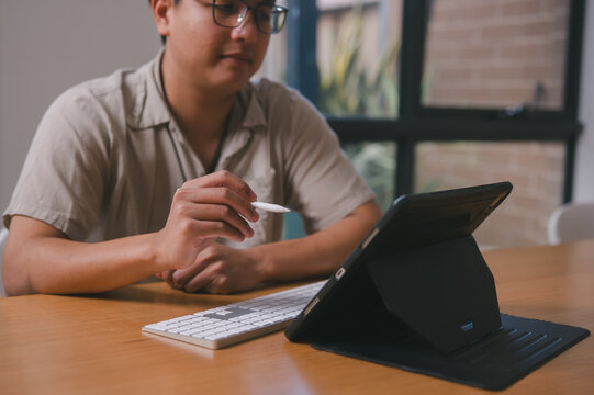 Young asian man working from home with digital tablet sitting in home office or living room with natural light, remote working data analysis and technology