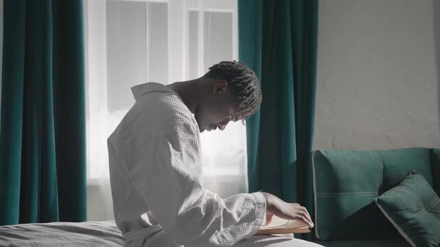 Guy in bathrobe sitting on bed, focused on reading book in soft natural light. Relaxing indoor scene with calm atmosphere, green curtains, and cozy environment. Quiet, peaceful moment