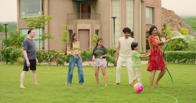 Indian happy mom man hold hose pipe spray water on house lawn park tree young adult family teen girl dad cute son kid boy child male female group play ball game have fun joy spend time enjoy trip day