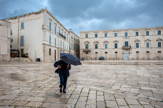 Un uomo regge un ombrello nero contro il vento forte nella spaziosa piazza  di Trani, delimitata da storici edifici in pietra. Il cielo coperto suggerisce un tempo inclemente.