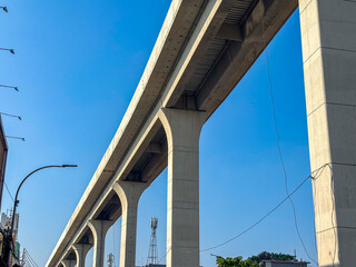 An elevated concrete LRT Jakarta track, under construction, rises on massive pillars against a clear blue sky. Visible red rebar framework highlights ongoing urban infrastructure development and progr