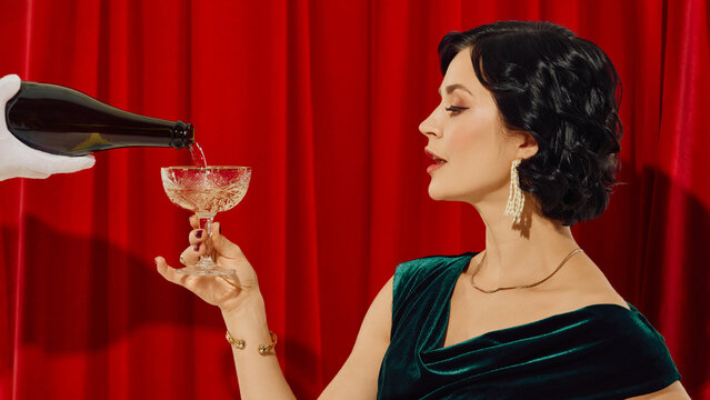 Woman in velvet dress holding champagne glass while bottle pours in front of red curtain. Concept of celebration glamour, luxury branding, sparkling drink ads, elegant lifestyle.