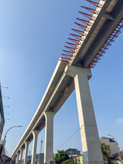 An elevated concrete LRT Jakarta track, under construction, rises on massive pillars against a clear blue sky. Visible red rebar framework highlights ongoing urban infrastructure development and progr