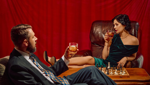 Elegant man and woman in formal wear sipping whiskey near chessboard in red-curtained room. Concept of refined leisure, intellectual lifestyle, luxury beverage ads, retro storytelling.