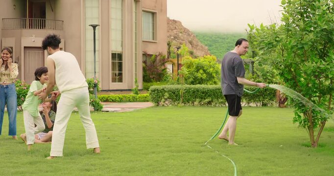 Indian happy dad man hold hose pipe spray water on outdoor house lawn park tree young adult family teen girl cute son kid boy child male female group play ball game have fun joy spend time enjoy trip