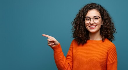 Smiling young woman with curly hair wearing glasses and an orange sweater points to the left against a blue background