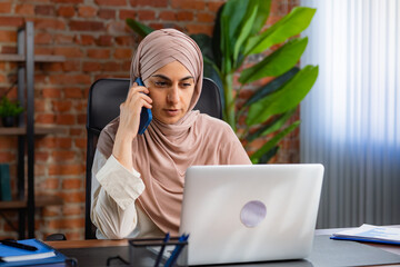Focused Professional Woman in Hijab Using Phone and Laptop at Office Desk