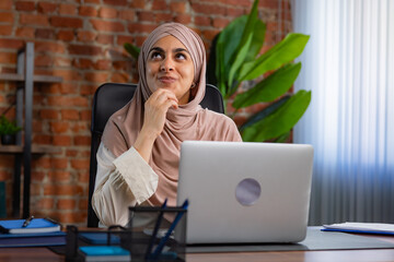 Middle Eastern Muslim woman in beige hijab working on her laptop and thinking about something.