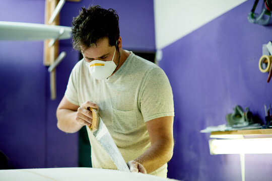 Man, saw and craftsman with foam, surfboard and PPE for skills, talent or hobby safety. Creative person, artist or entrepreneur with workshop for design manufacturing or custom work for artisan craft