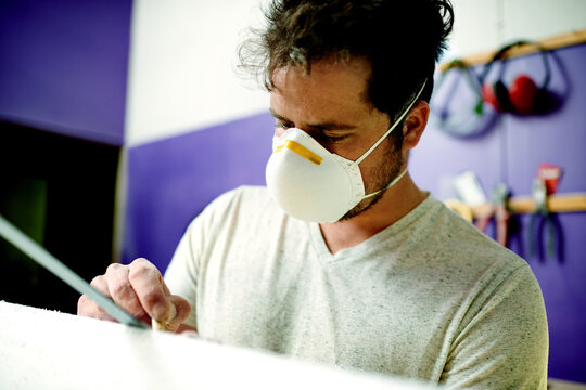 Manufacturing, man and sanding surfboard in workshop for production process, design or creative craftsmanship. Surfer, blank or person with tools for shape, polish or smooth foam with protective mask