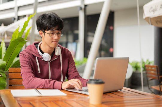 Glasses asian man or student looking typing on laptop while sits with coffee at wooden table in cafe