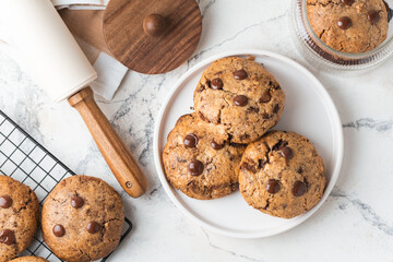 Fresh chocolate American chip cookies with nuts and chocolate drops on white marble background. Flat lay of cookies with stylish cutlery and pastry chef accessories. Recipe of homemade cookies concept