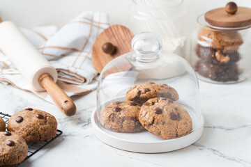 Stack of fresh chocolate American style chip cookies with nuts and chocolate drops on a tray with transparent lid on white marble background. Cookies inside a glass jar with kitchen utensils
