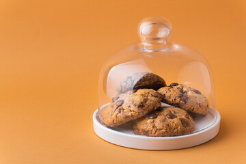 Stack of fresh chocolate American style chip cookies with nuts and chocolate drops on a tray with transparent lid on warm brown background. Cookies with peanut butter filling inside a glass jar