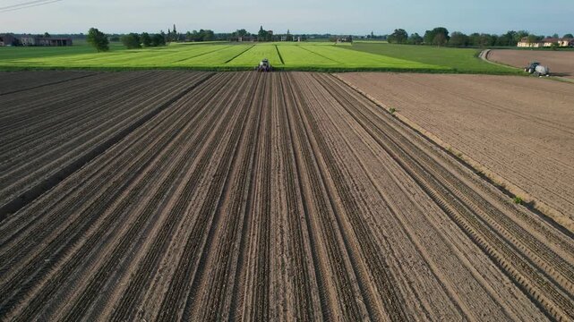 Expansive drone view shows farm tractors and agricultural workers efficiently sowing tomato plants on neat, harrowed fields surrounded by patchwork crops, slow motion shot