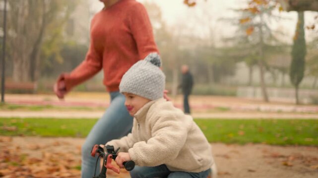Mother encouraging child riding bike in autumn park path.