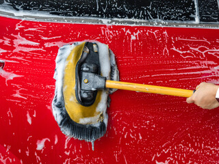 Close-up of a person holding a mop washing a red car and cleaning it with foam at a self-service car wash. © Pornpimon