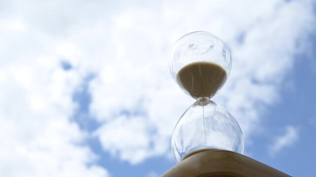Glass hourglass against blue sky with clouds. Transparent hourglass filled with sand placed against a cloudy blue sky background. Concept of time, transience, waiting, and the passing of life - Powered by Adobe