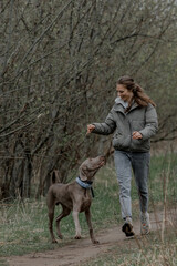 A young Caucasian woman with brown hair runs joyfully on a dirt path with a playful brown dog in a green, wooded area.
