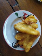 A close-up of golden fried bananas, a traditional Indonesian fried banana snack. The crispy fritters are served on a classic rooster plate, set against a rustic wooden table.