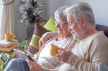 A modern, smiling older couple relaxes at home, sitting comfortably on the couch, surfing the...
