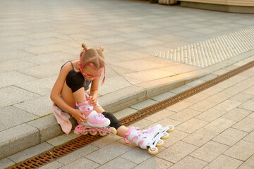 Focused young girl in black top and denim shorts sitting on curb at sunset, fastening her roller skate, capturing a quiet moment of childhood concentration outdoors