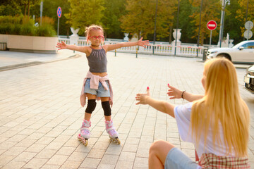 Joyful girl in gray top and denim shorts skating outdoors during golden hour, arms spread wide and smiling, capturing childhood happiness in a lively city park scene