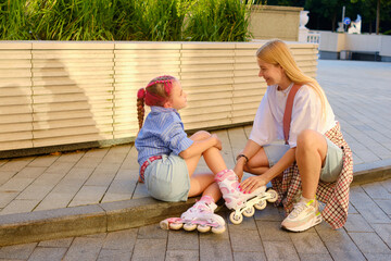 Joyful young woman helping cheerful girl with pink hair put on roller skates outdoors on sunny day, vibrant urban park setting capturing playful lifestyle moment