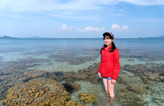 Young Asian girl wearing a red UV-protective long-sleeved shirt and cap stands in the shallow ocean surrounded by coral reefs on a sunny day, smiling at the camera. Koh Kradan, Trang, Thailand.