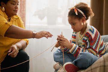 Young Black girl learning knitting while spending quality time with grandmother in living room, intergenerational relationship concept