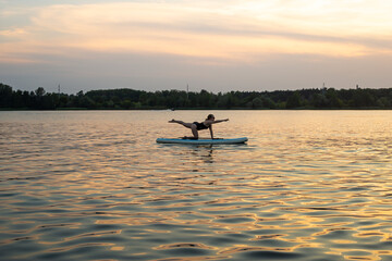 Naklejka premium woman doing yoga on sup board at sunset. outdoor summer activity. Sup yoga. Social Distancing. copy space. Mental Health