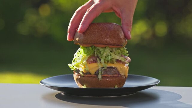 cook put bread bun on freshly grilled burger meat with melted cheese and onions on bread bun with sauce on plate in sunny garden, food closeup, outdoor cooking