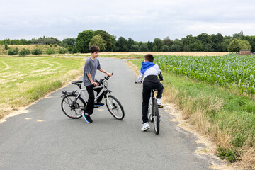 Obraz premium Two boys riding bicycles on a rural road surrounded by green fields and trees. 
